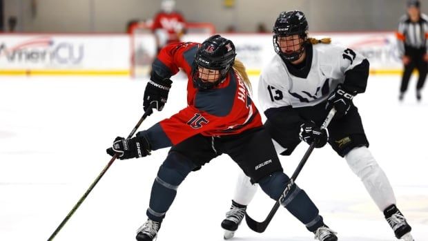 A female hockey player in a red jersey, with number 15 on the side of her sweater, keeps the puck away from another female hockey player in a white and black jersey.