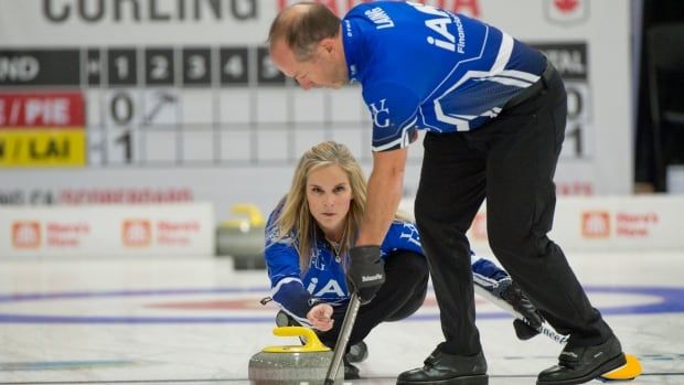 A female curling skip delivers a stone with her right hand as he male mixed doubles partner sweeps ahead of her.