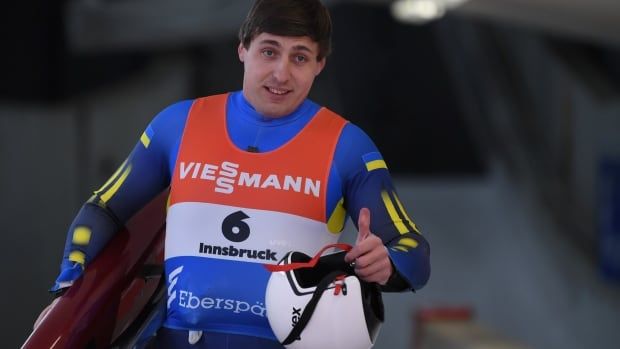 A luge athlete flashes a thumbs up and smile as he carries his sled.