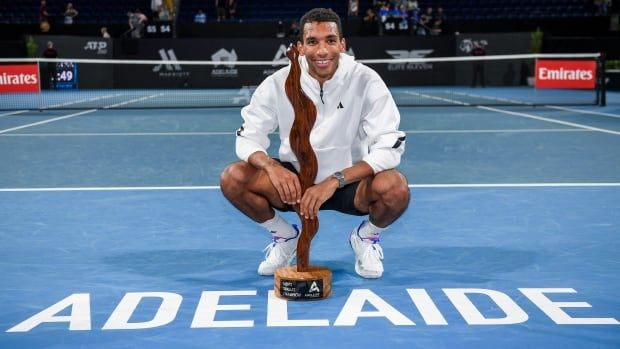 A men's tennis player squats on a blue tennis court in front of a tall and thin wooden trophy.