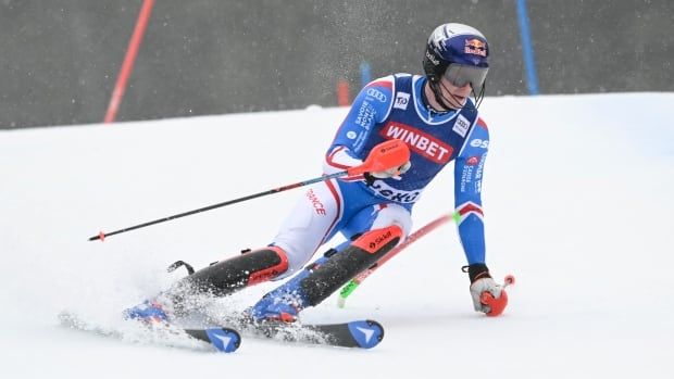 A men's skier, wearing a light blue and white ski suit with a dark blue bib, navigates around a gate on a ski hill.