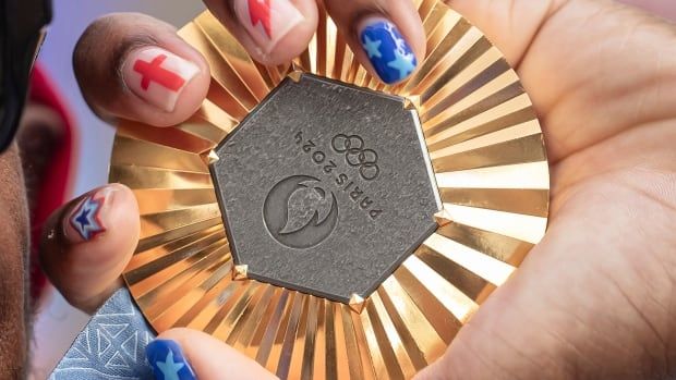 A gold medal is shown being held by a hand with painted fingernails.