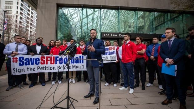 Man in a suit speaks in front of microphone to a crowd of people, some of them holding signs that read 'Save SFU Football.'