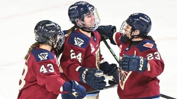 Three female ice hockey players smile while wrapping their arms around each other in celebration on the ice during a game.