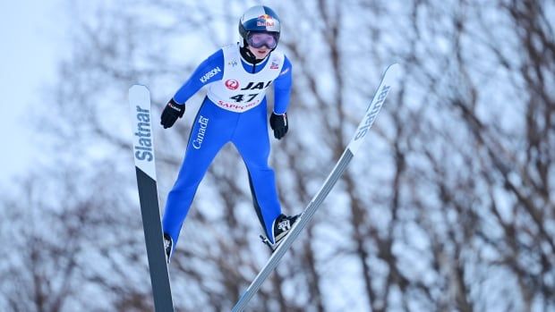 A female ski jumper soars through the air with trees in the background during the day.