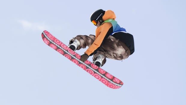 A male snowboarder flies through the air while grabbing his board with his left hand during a daytime slopestyle event.