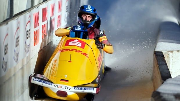 A German four-man bobsleigh crew speeds down a track as snow flies behind them.