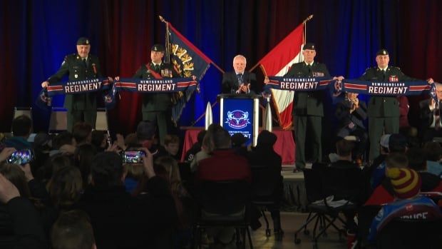 Four men in military uniform stand on a stage holding navy blue and red scarves. The scarves read 'Newfoundland Regiment'. A man wearing a suit stands at a podium in between them.