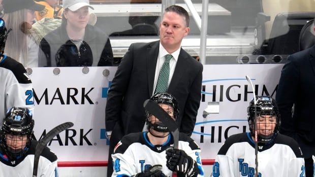 A man in a suit stands behind several hockey players on a bench.