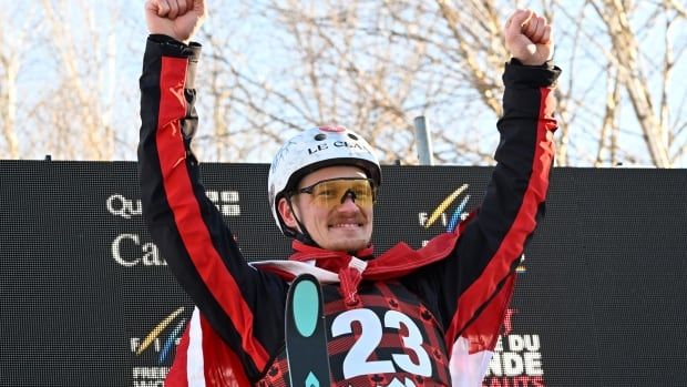 A male aerials skier smiles while raising both arms in celebration on a podium.