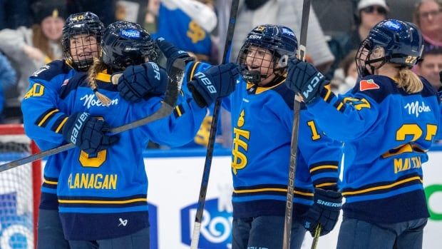 Four female ice hockey players smile while wrapping their arms around each other in celebration during a game as fans watch from behind the glass.