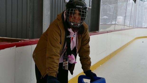 A girl wearing a hockey helmet smiles while holding onto a blue skating glider at an ice rink.