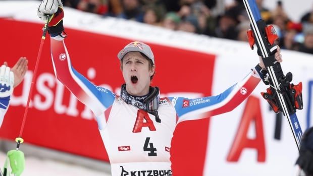 A male alpine skier raises his arms while shouting in celebration at the end of a slalom course as fans watch from behind a barricade.