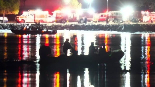 In an nighttime scene, a patrol boat with a few people on board is barely visible on a body of water, with multicolored lights seen from the shore to illuminate the scene.