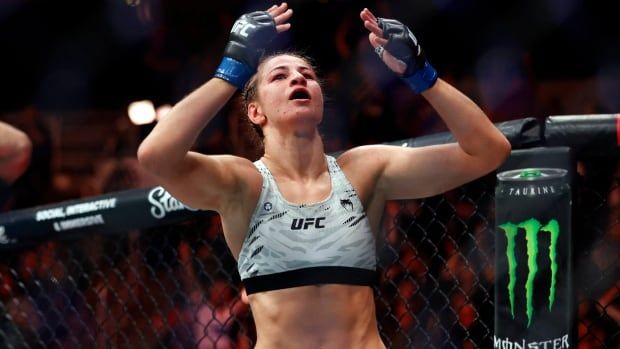 A female mixed martial artist raises her arms in celebration inside a UFC octagon as fans cheer in the background.