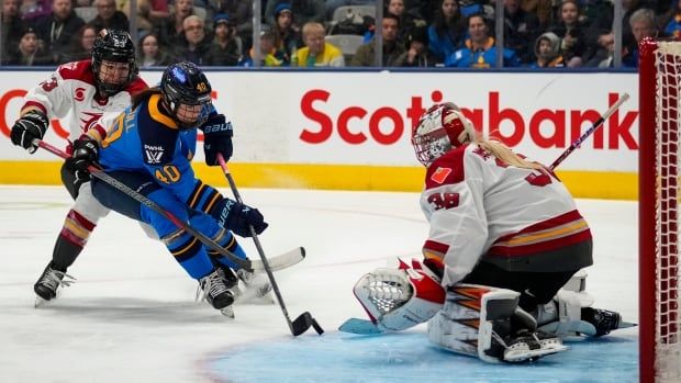 A female hockey player shoots on a goaltender from close range as a defender hooks her while fans watch from behind the glass.