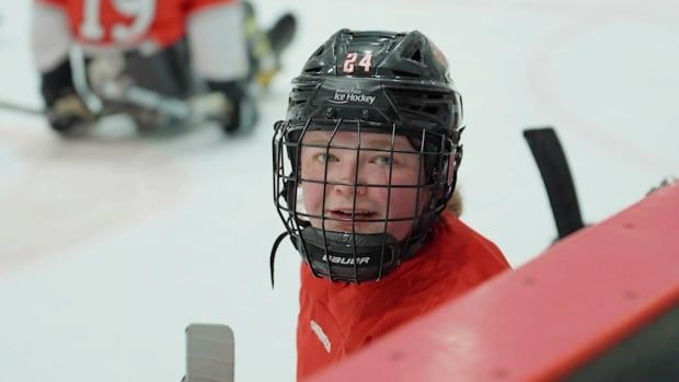 A women wearing a black hockey helmet and red hockey jersey looks up over the sideboards at a hockey arena. There is a sledge hockey player on the ice in the background.