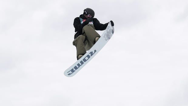 A male slopestyle snowboarder soars through the air while holding the tip of his snowboard with his left hand during a daytime competition.