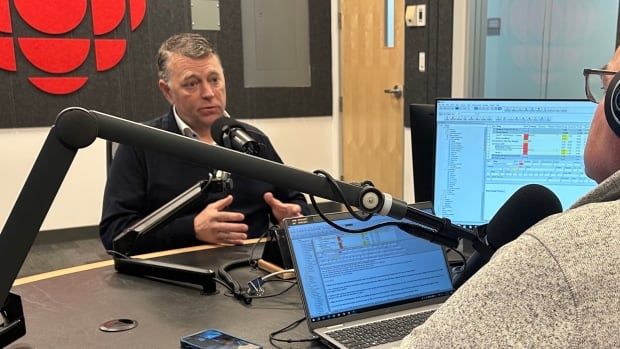 Two men seated at a table, with a laptop on a monitor on the table, engaged in conversation in a studio with microphones positioned between them