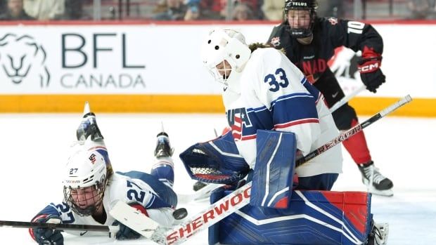 A women's hockey goaltenderkneels in butterfly position as her teammate dives in front of her. An opposing team player is shown in the background.