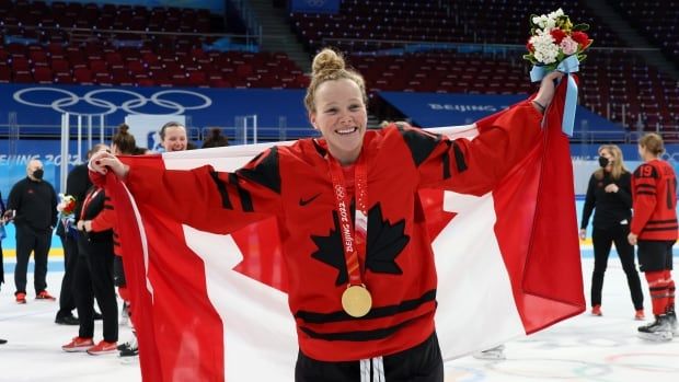 A hockey player in a Canada jersey celebrates with a gold medal around her neck while holding the Canadian flag.