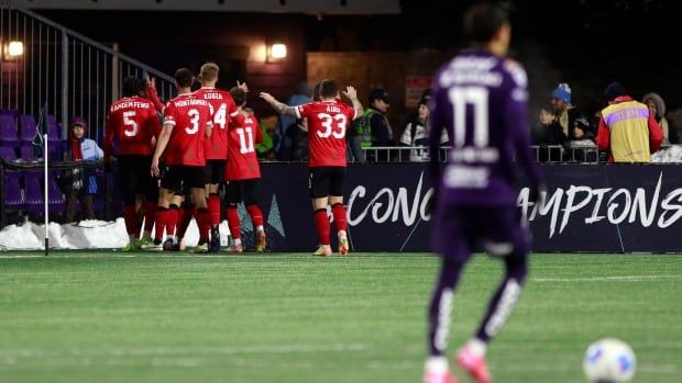 A soccer team is pictured celebrating a goal in the background with a blurred opposition team member pictured in the foreground.