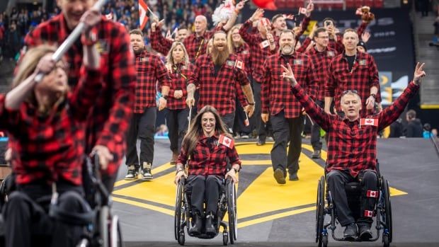 Athletes in plaid jackets parading, some in wheelchairs, waving the Canadian flag. 