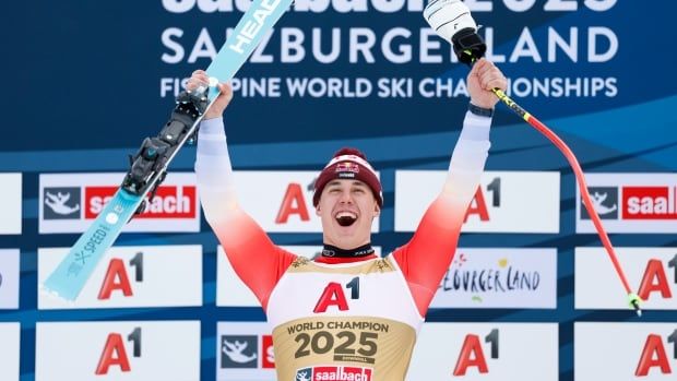 A male alpine skier smiles on the podium while lifting his skis and poles into the air.