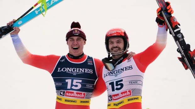 Two men's skiers put an arm on one another and raise their skis in celebration.