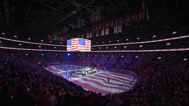 Hockey players stand in a rink as the U.S. flag is projected on the ice and a large monitor above them.