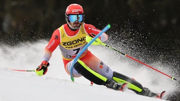 A male alpine skier races down a slalom course as snow flies into the air around him.