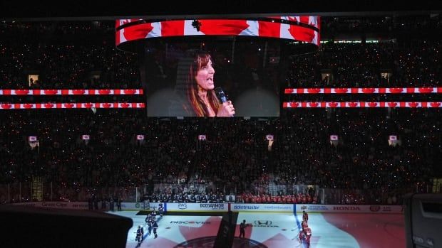 A singer with brown hair is seen on a Jumbotron inside a hockey arena with Canadian flags.