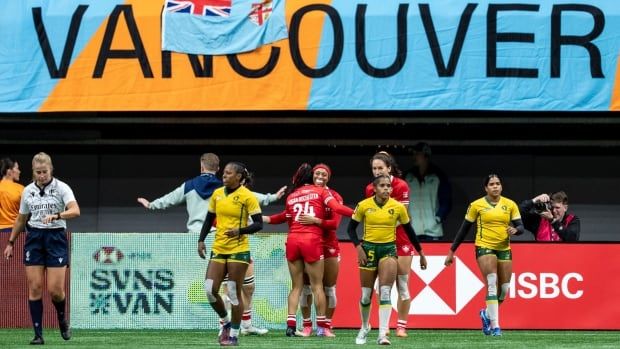 Several women's rugby players stand on the pitch after a try. A large flag above the pitch says 'VANCOUVER'