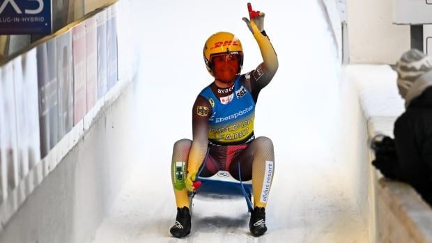 Female luge athlete sits up on sled after finishing race, points a finger in air to celebrate. 