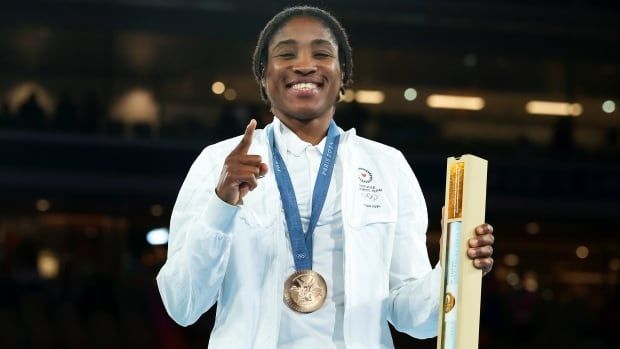 A female boxer with an Olympic medal around her neck smiles while holding up her right index finger.