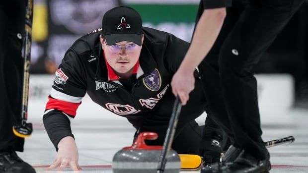 A male curling skip wearing glasses crouches down with his right hand on the ice as he watches his teammates sweeping ahead of a stone.