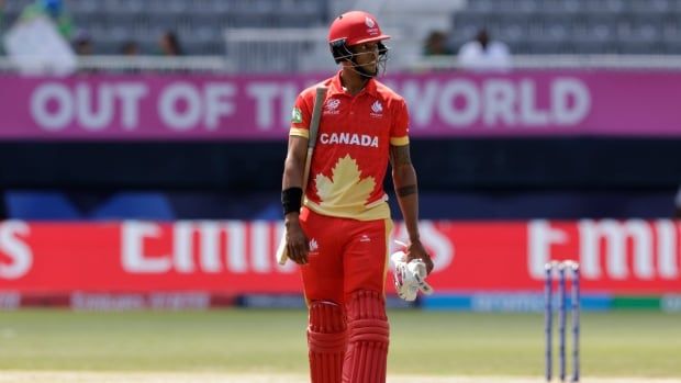 Male cricket player walks across cricket field with hitting gloves and bat in hand.