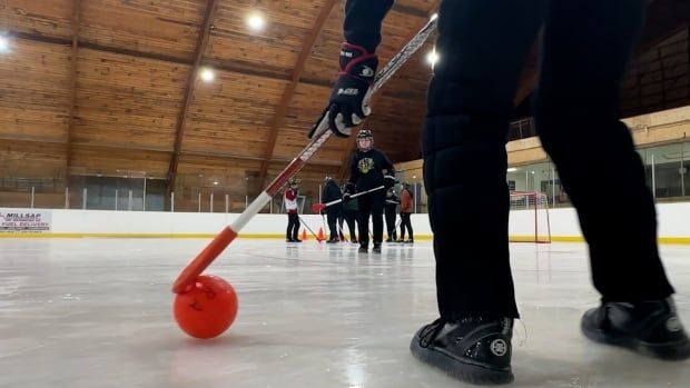 A long angle on an indoor ice rink of a young person stick-handling an orange ball with a broom.