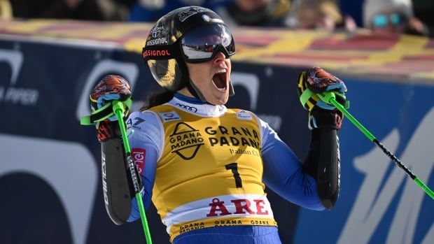 A female alpine skier raises her poles as she yells in celebration at the bottom of a course.