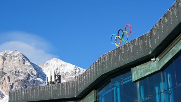 A building with the Olympic rings on it is shown, with the mountains in Italy as a backdrop.