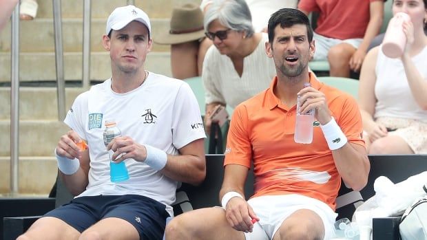 Two men's tennis players sit on the sideline holding drinks.