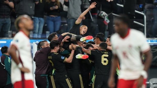 A group of men's soccer players huddle together while one jumps on top of them. Two opposing players are in the foreground of the photo, unfocused.