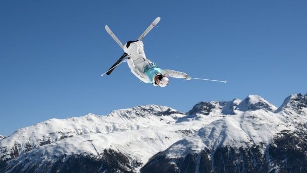 Male freestyle skier goes airborne, pictured upside down with skis crossed, with mountains in background.